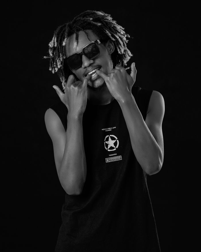Black and white portrait of a stylish young man with dreadlocks and sunglasses striking a confident pose.